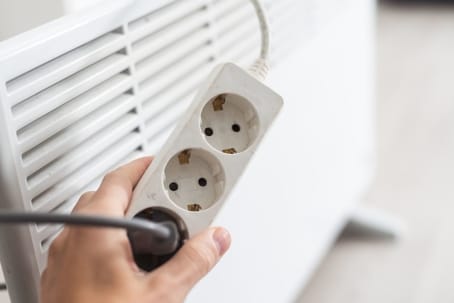 Hand holding a power strip with multiple outlets, positioned near an air conditioning unit, emphasizing the importance of proper electrical connections for HVAC maintenance.