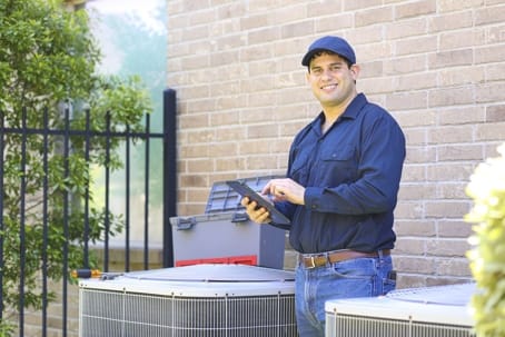 A technician in blue work clothes uses a tablet next to air conditioning units outside a building, ensuring top-notch AC maintenance and reliable local HVAC repair services.