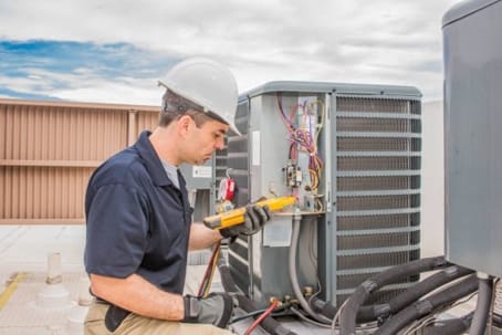 AC technician wearing a helmet and gloves uses a multimeter to check an HVAC system on a rooftop during routine ac maintenance.