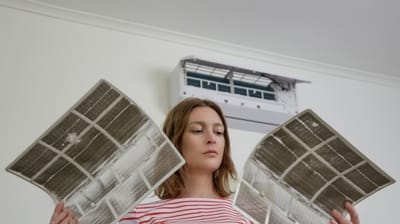 Woman holding air conditioner filters, examining them, with a wall-mounted air conditioning unit in the background, related to HVAC maintenance and efficiency.