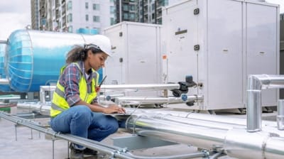 Engineer in a hard hat and safety vest working on a laptop near industrial pipes and machinery on a rooftop in California.