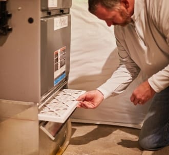 An AC technician in a white shirt is changing the air filter of a home furnace as part of routine AC maintenance.